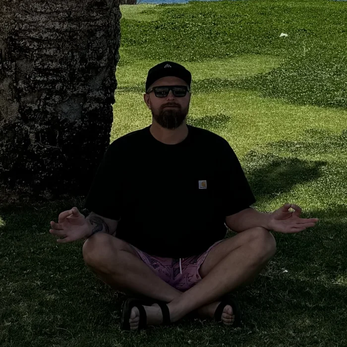 André meditating cross-legged on the grass under a palm tree, the ocean behind him